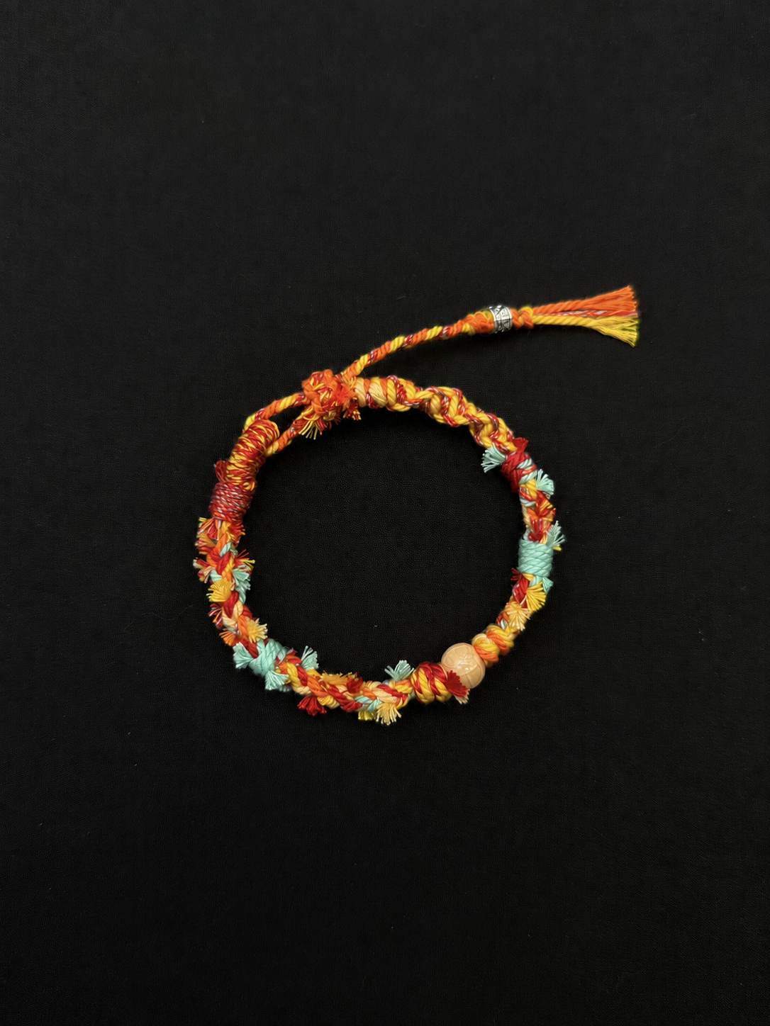 Apricot Breeze orange, yellow and mint woven bracelet with a peach-toned round bead and a compact braided tassel. Product view on dark fabric.