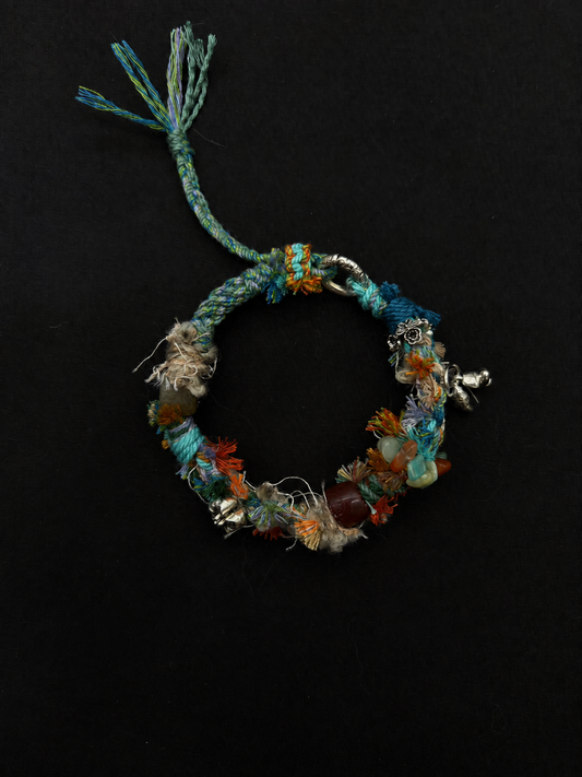 Cove Echo teal, orange and cream woven bracelet with textured knots, mixed decorative beads and small dangling details. Product view on dark fabric.