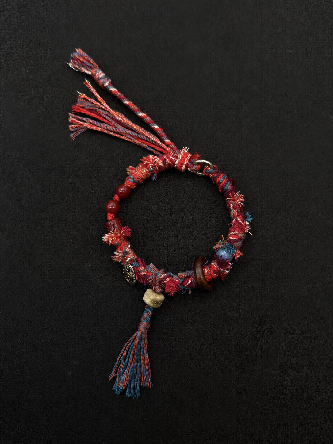 Dusk Ember red, blue and cream woven bracelet with long red-and-blue tassels, round beads and a small disc charm. Product view on dark fabric.