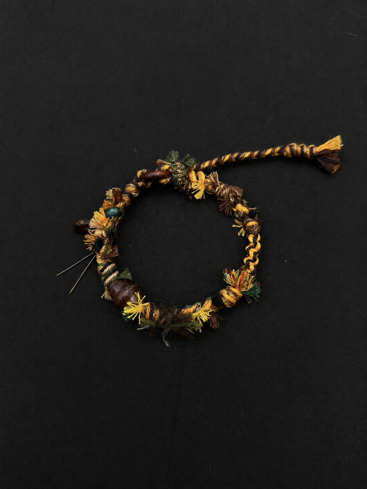 Earthbound brown, mustard and olive woven bracelet with earth-toned decorative beads and a compact braided cord end. Product view on dark fabric.