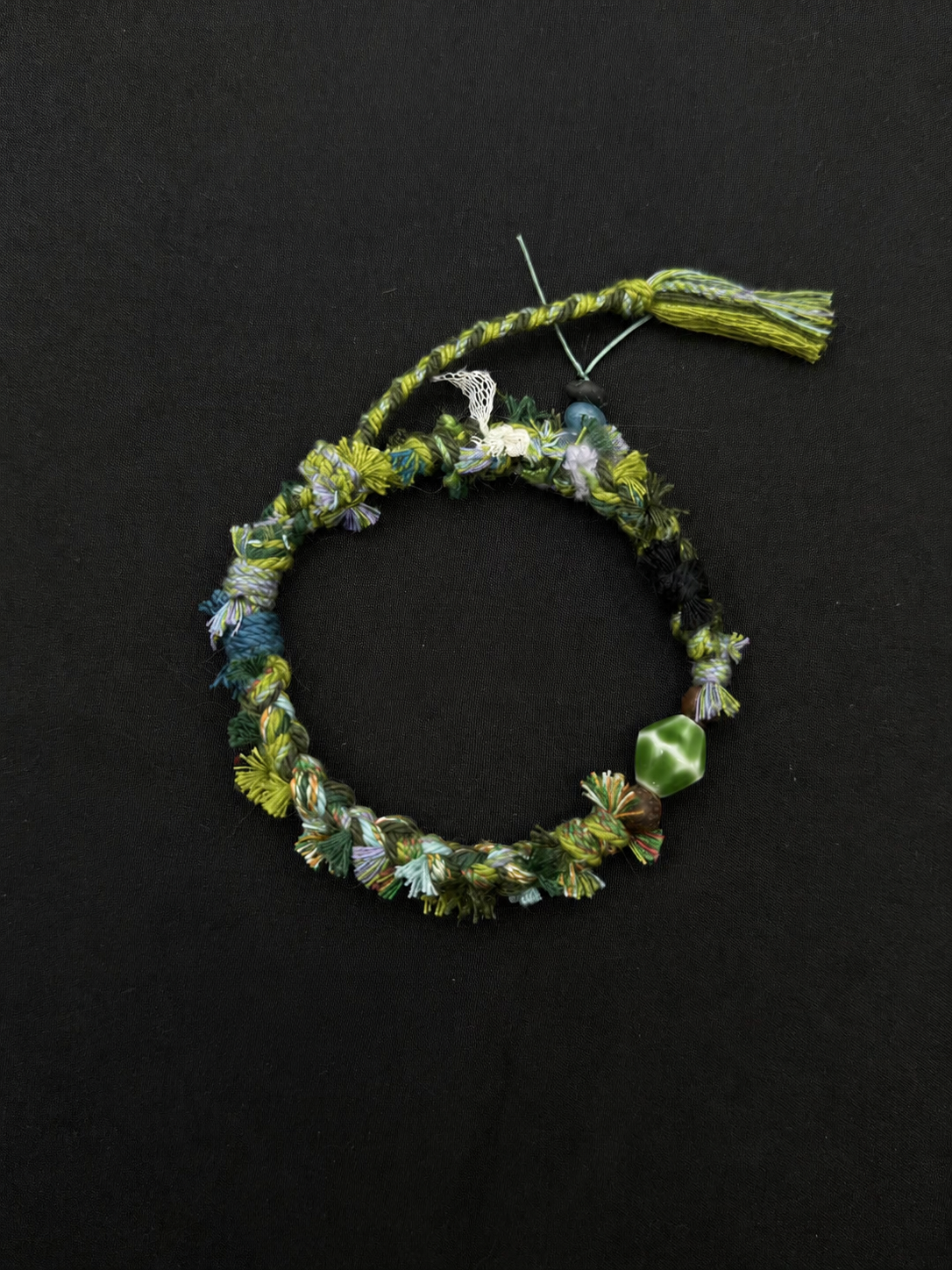 Fern Passage green, blue-gray and charcoal woven bracelet with a faceted green accent bead and softly textured cord. Product view on dark fabric.