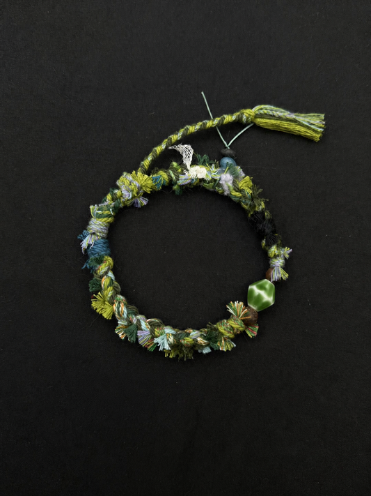 Fern Passage green, blue-gray and charcoal woven bracelet with a faceted green accent bead and softly textured cord. Product view on dark fabric.