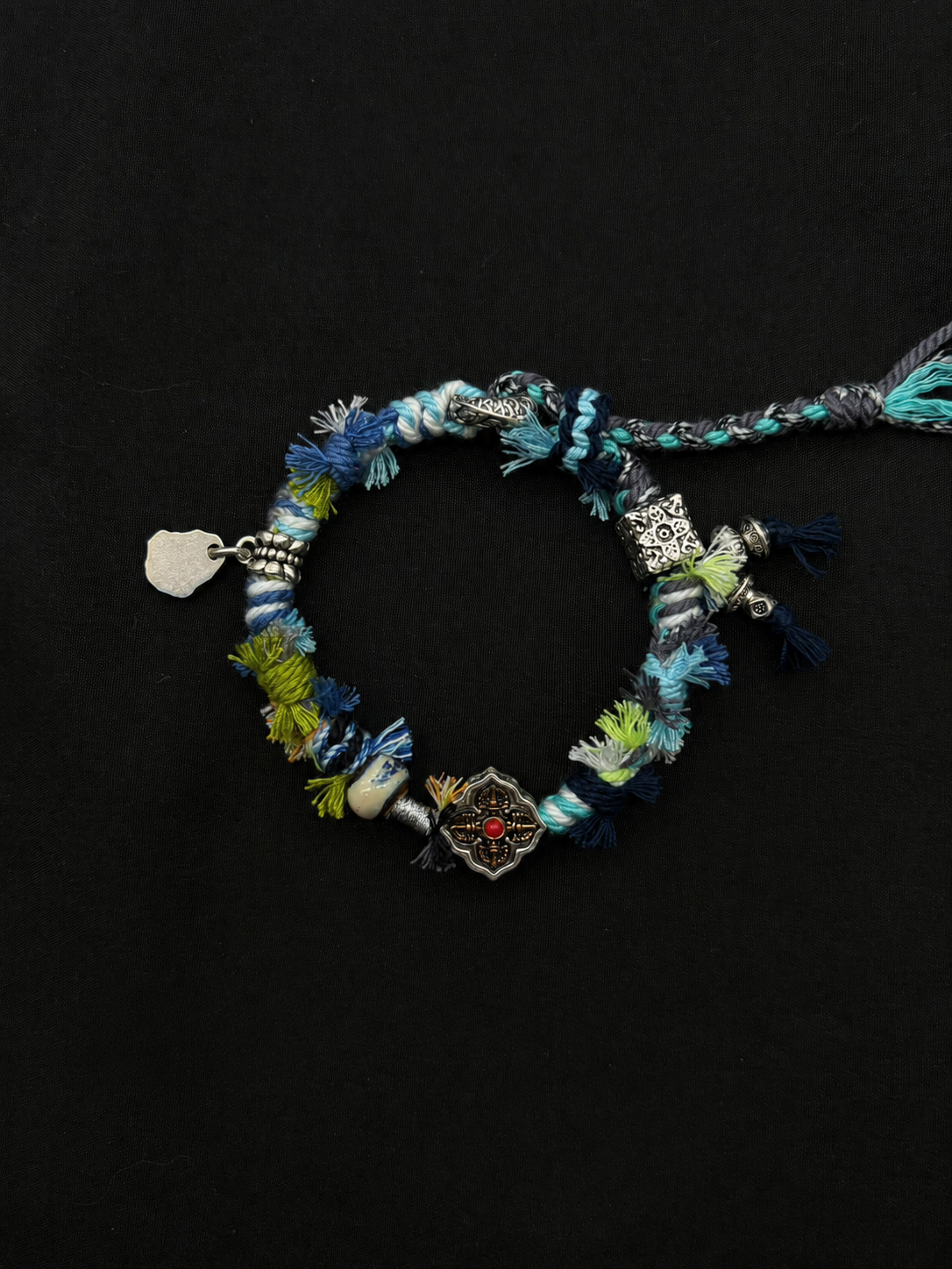 Meadow Current blue, green and turquoise woven bracelet with a flower-shaped centerpiece with a red accent and silver-tone details. Product view on dark fabric.
