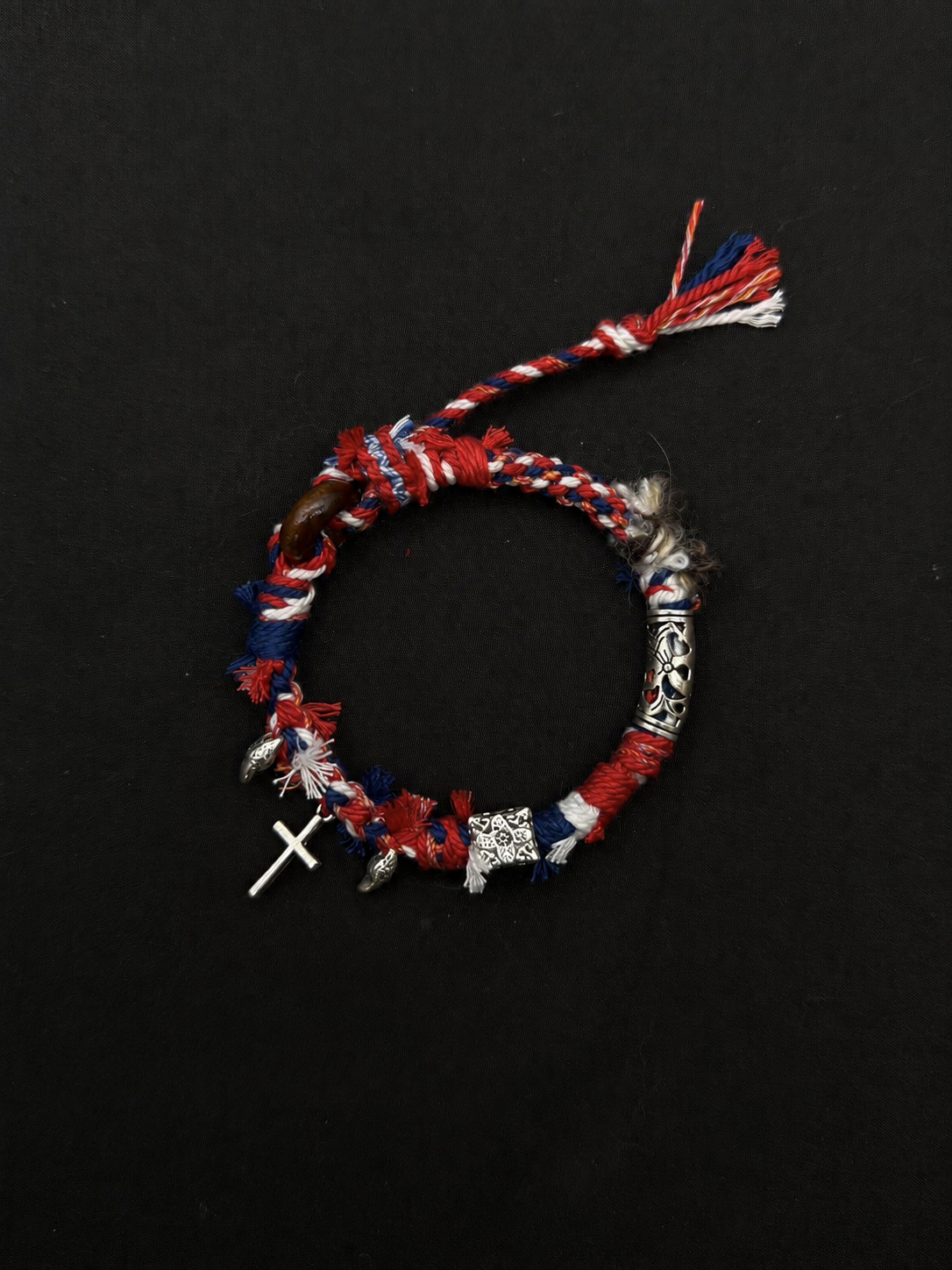 Northward red, navy and white woven bracelet with a cross-shaped charm and patterned silver-tone tube accent. Product view on dark fabric.