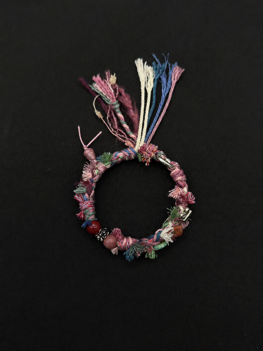 Rosewind pink multicolor woven tassel bracelet with decorative beads, shown on a dark fabric background.