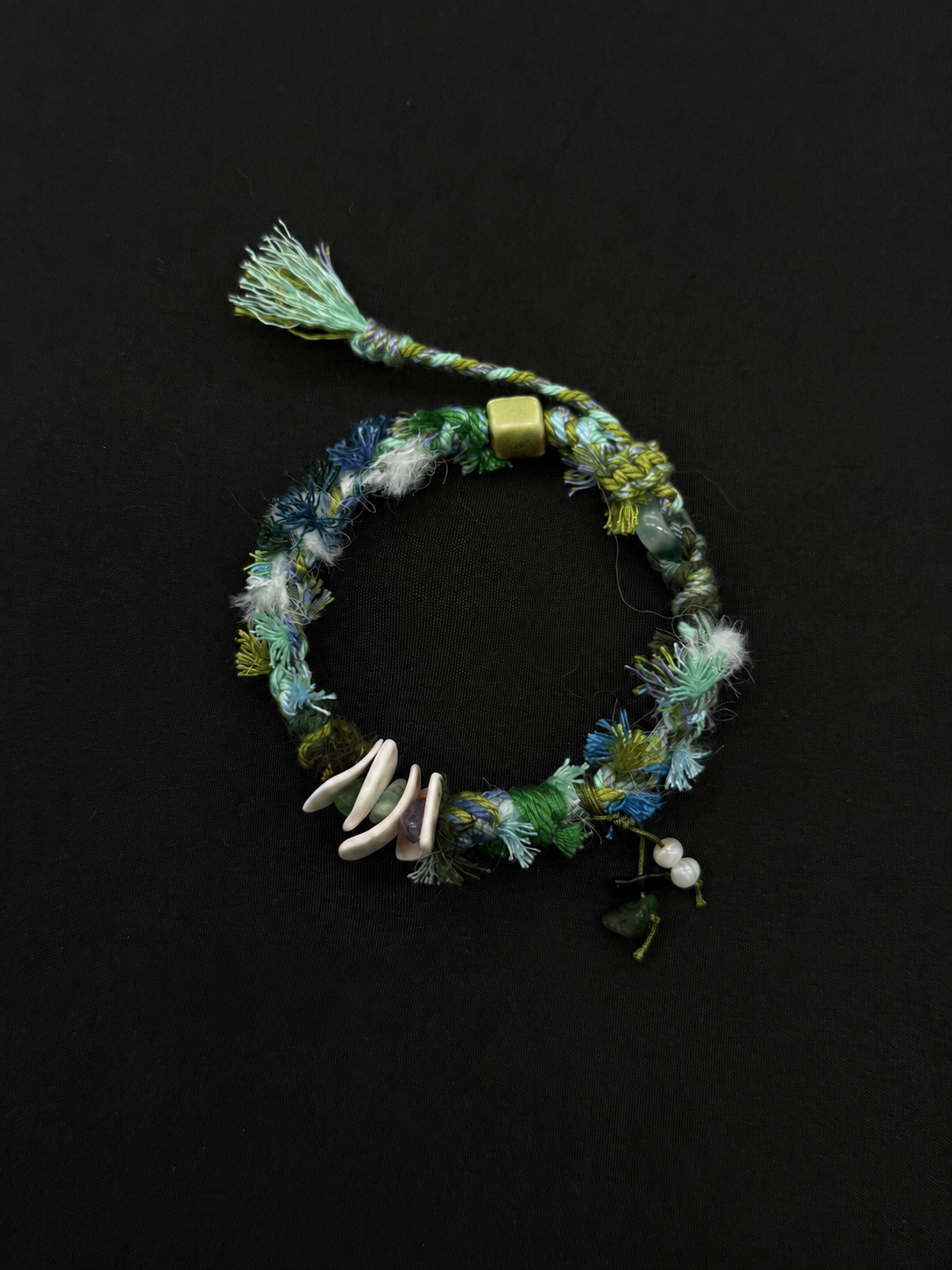 Seafoam teal, green and blue woven bracelet with white curved accents, textured cord and a short tassel. Product view on dark fabric.