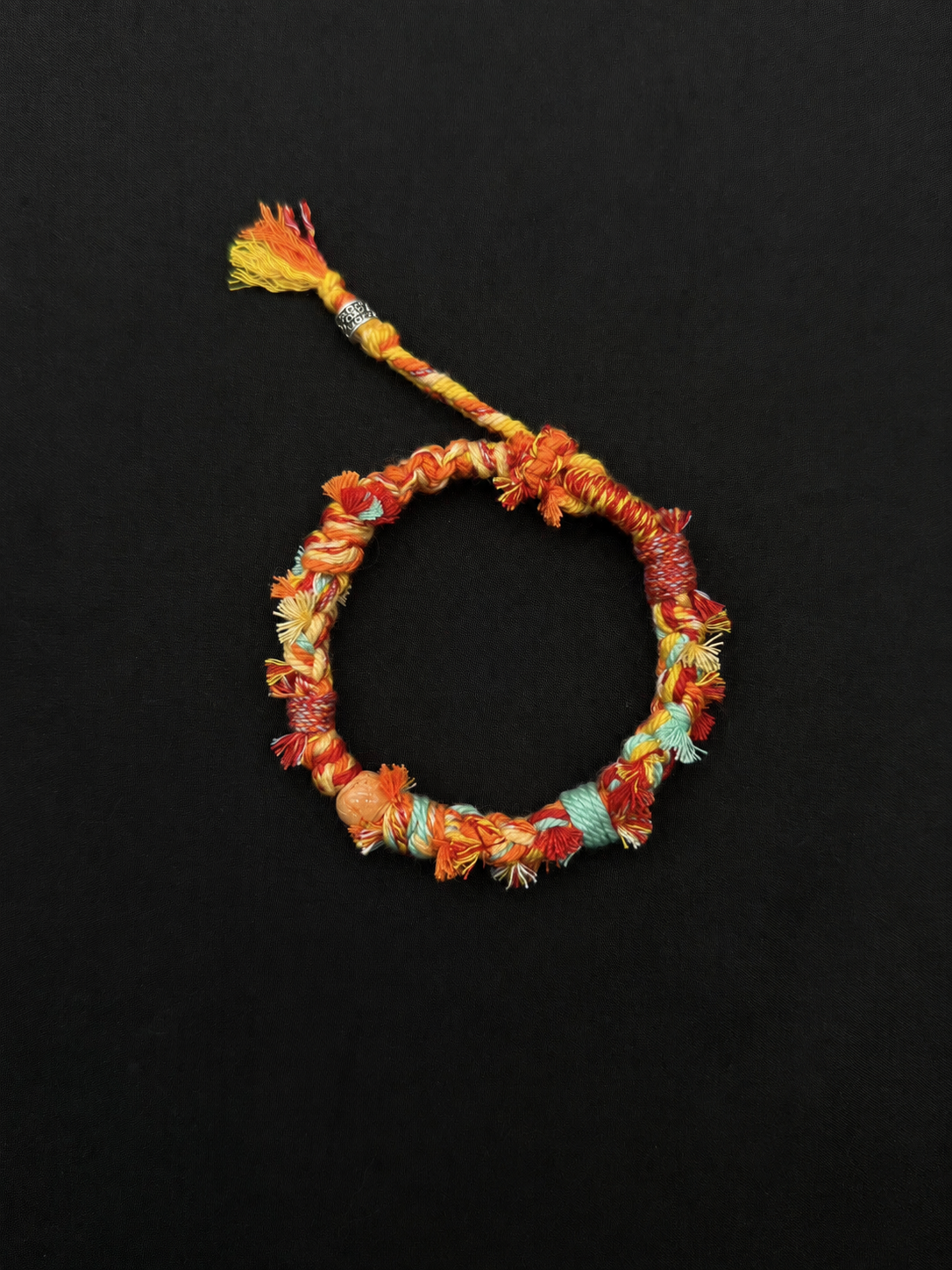 Sundrift orange, yellow and pale turquoise woven bracelet with warm woven color, a peach-toned bead and a short tassel. Product view on dark fabric.