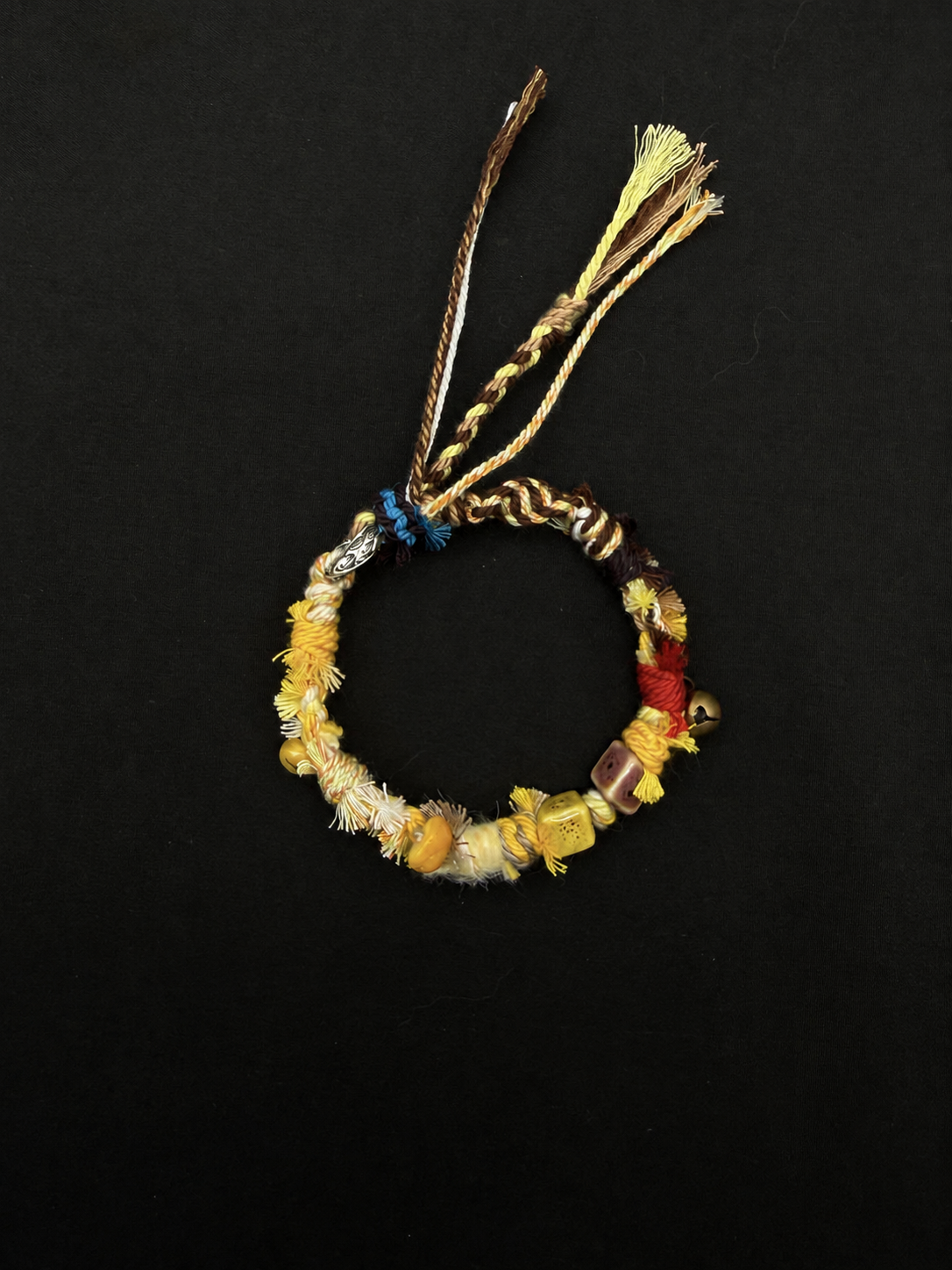 Sunlit Dune yellow, cream and brown woven bracelet with square decorative beads, a small bell detail and long cord ends. Product view on dark fabric.