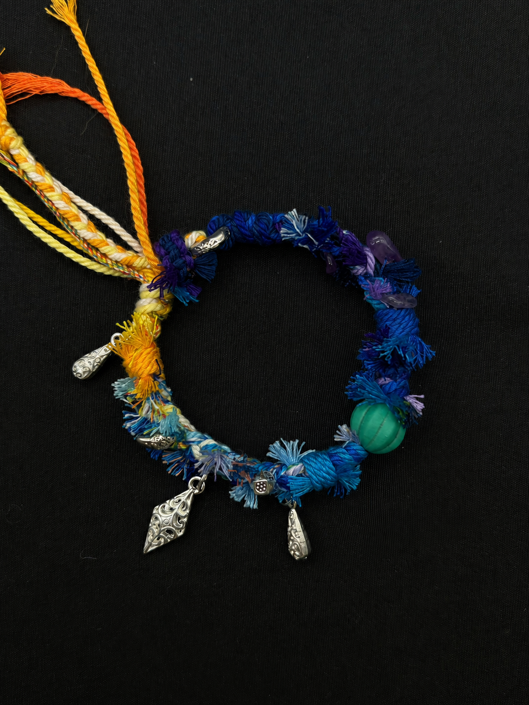 Twilight Tide blue, violet and yellow woven bracelet with a grooved green bead, silver-tone drops and bright cord ends. Product view on dark fabric.