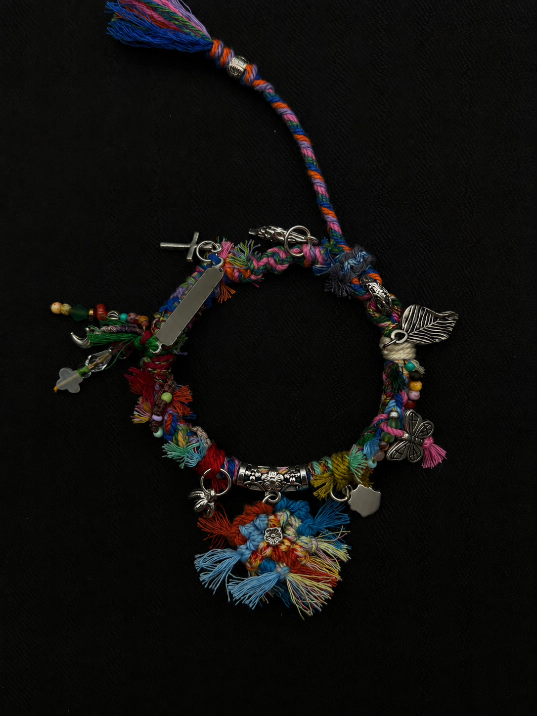 Wildflower Drift blue, pink, green and red woven bracelet with a clustered tassel flower and an assortment of silver-tone charms. Product view on dark fabric.